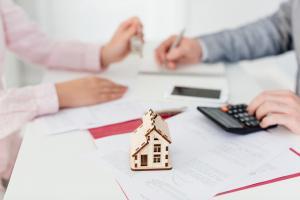 Mortgage Refinance Graphic shows a wooden home sitting on a table. Behind the home are 2 people facing each other, one with keys in their hand and the other with a calculator and pen.